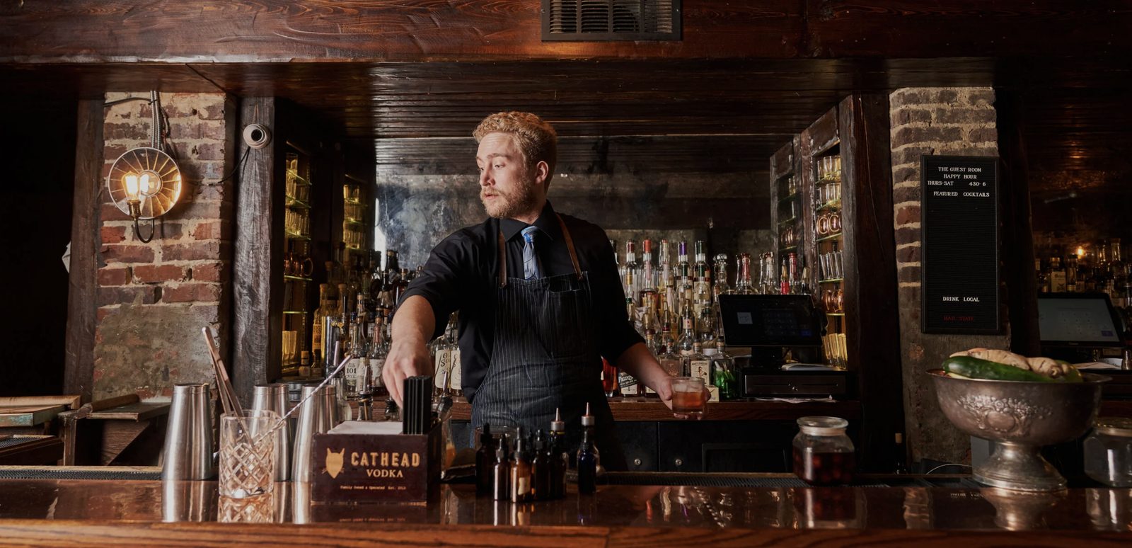 A bartender reaching across the bar at The Guest Room, with brick walls, warm lamps and shelves of spirits behind him
