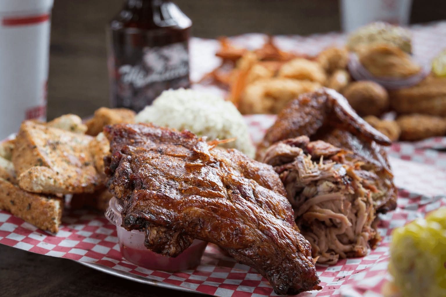 Smoked ribs and pulled pork with coleslaw and toast on red-checked paper, a bottle of Little Dooey barbecue sauce behind